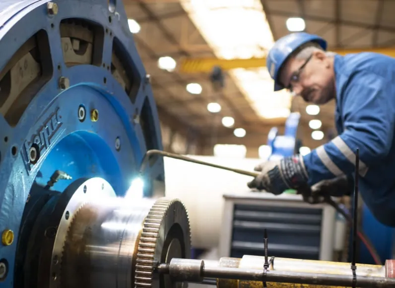 In a workshop, a technician repairs blue drilling equipment with welding and metal machining, supported by API-certified workshops in Europe and Middle East.