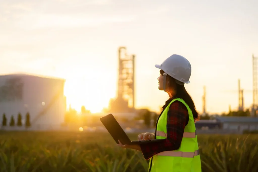 Exuocova governance scene: a female worker in a white hard hat and bright yellow safety vest with a clipboard stands at an industrial site during sunset, conveying safety and compliance.