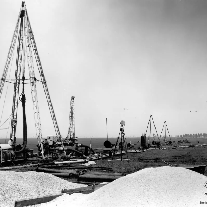 Historic black-and-white photograph of early drilling rigs in an expansive oilfield, featuring tall derricks and industrial equipment associated with EXUOCOVA heritage era.